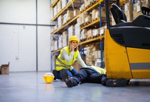man laying on ground while coworker calls for help after forklift accident in warehouse